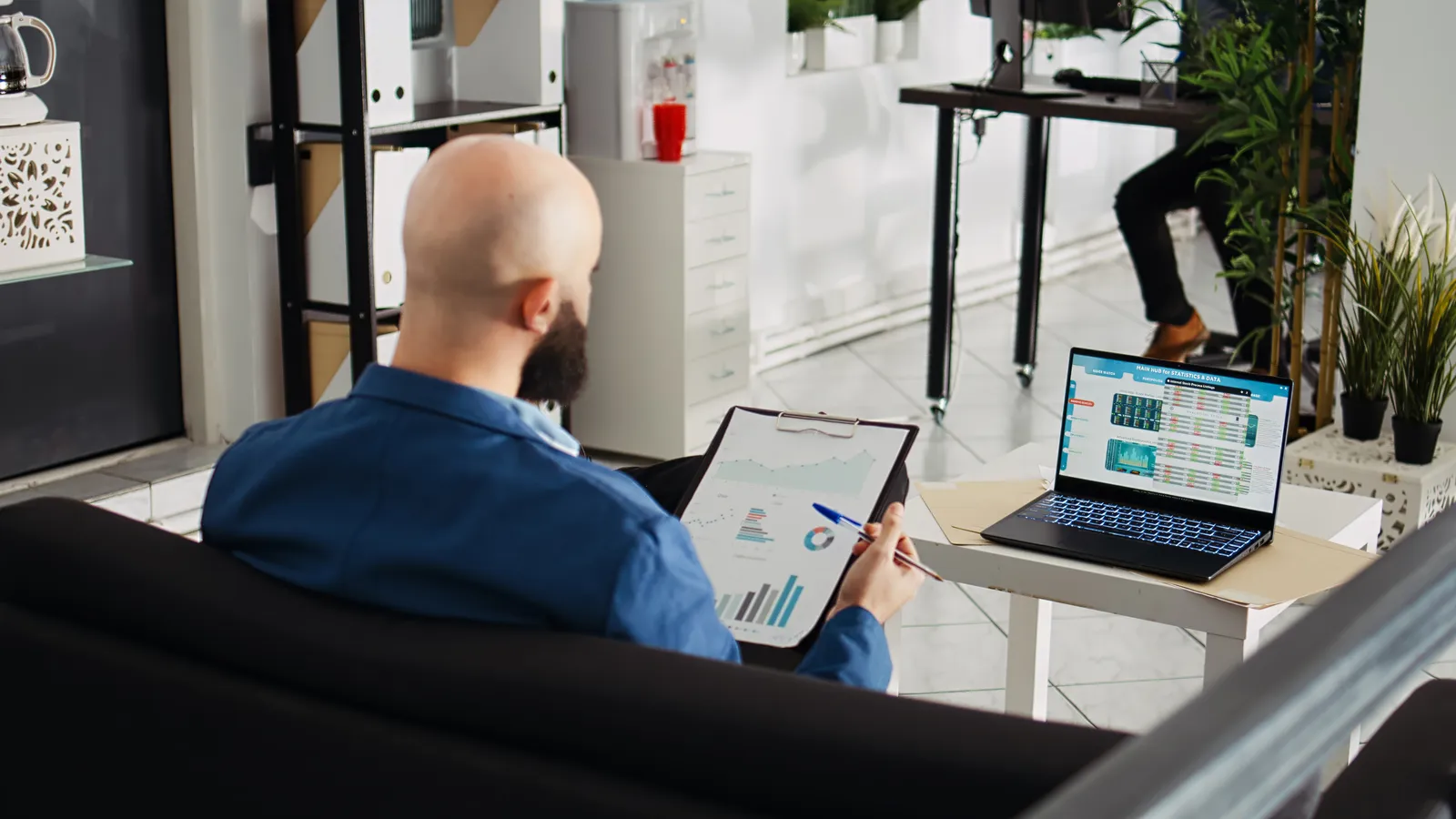 A man reviews a tablet and laptop with charts at a small office